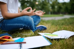Cropped view of a school child boy sitting in lotus position on green grass of city park and meditating . Workbooks school supplies lying on the grass. Concentration, recreation, mindfulness concepts