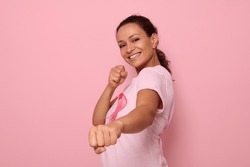 Pretty woman in pink t-shirt and cancer awareness ribbon stands in a fighting stance to mark the fight against cancer, in honor of October 1, smiles looking at camera, colored background, copy space