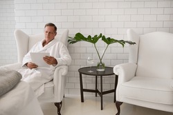 50 years old business man wearing white terry bathrobe reading price list of catalog of beauty procedures sitting on an armchair of a luxury white wellness spa clinic