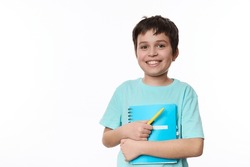 Lovely primary school student, smart teen boy in blue casual tshirt, holding textbook and yellow pen, smiling a cute toothy smile looking at camera, isolated, white background. Copy advertising space