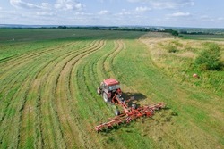 A red tractor rakes the mown grass for drying. Modern equipment on the field. Preparations of fodder grasses for hay. Top view.