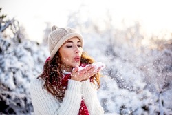 Happy woman walking in the snowy winter day outdoor. Female mdel dressed white sweater and hat blowing snow and posing against the  sun 