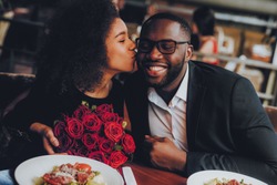 African American Couple Dating in Restaurant. Romantic Couple in Love Dating. Cutel Man and Girl in a Restaurant Making Order. Romantic Concept. Girl Kissing Man. Bouquet of Flowers. Red Roses.
