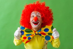 Portrait of a funny playful clown in red wig showing his big colorful bow tie, looking at camera and smiling, on a green background