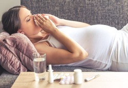 Side view of beautiful pregnant woman having cold, wiping her nose while lying on couch at home. Medicine is on the table