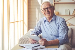 Handsome old man in eyeglasses is using a digital tablet, looking at camera and smiling while working at home