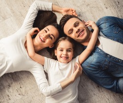 Top view of cute little girl and her beautiful young parents looking at camera and smiling while lying on the floor at home