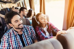 The guy and the girl are on the bus. The girl has headphones. He and the guy are discussing something and smiling. Behind them sit other tourists who are looking out the window to the sights.