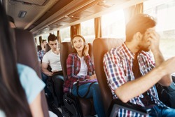 The girl is sitting on the bus in headphones and listening to music. She smiles and enjoys music. Next to the bus are other people who also went on a trip.