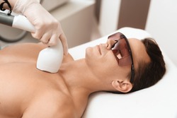 The man came to the procedure of laser hair removal. The doctor treats his neck and face with a special apparatus. The man has red glasses.
