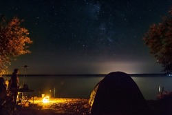 Silhouette of a male traveler near a tourist tent fishing. Fisherman resting on the shore of the lake near the fire and enjoying a beautiful view of the night sky, full of stars and the Milky Way