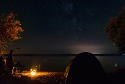 Silhouette of a male traveler near a tourist tent fishing. Fisherman resting on the shore of the lake near the fire and enjoying a beautiful view of the night sky, full of stars and the Milky Way