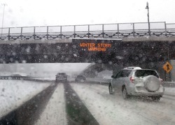 Winter Driving on Highway with Falling Snow on Windshield Passing Under a Bridge with an Electronic Winter Storm Warning Sign