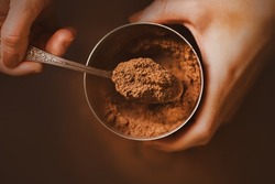A woman with a brown manicure on her nails holds a jar of fragrant crumbly cocoa powder in her hands and scoops a portion with a silver teaspoon. A delicious drink in the morning. The cooking process.