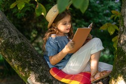 A cute little girl in a straw hat reading a book on a large tree. Children and reading. Concept of children's recreation without gadgets.