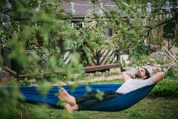 A man is resting in a hammock in a country house. A young guy lies in a hammock, dozing outdoors in the summer in the green of trees in the country