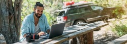 Male photographer working on a laptop outdoors in a camping. Young guy, freelancer, travel blogger working online at a laptop computer while hiking in the mountains outdoors. 