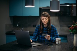A young woman is shopping online with a laptop at the table in her apartment. A beautiful brunette girl in a dress sits with a computer at a table in a stylish apartment, buys in an online store