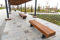 Empty park seats made of thick timber with metal legs set in a newly refurbished park. The top is covered with wood on a metal frame, providing protection from the sun.