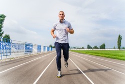 Full length front view on adult caucasian man jogging on the running track - Blonde male athlete in stadium training run in sunny spring or summer day - real people healthy lifestyle concept