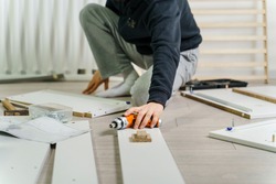 One unknown Man alone Putting Together Self Assembly Furniture sitting on the floor at Home holding electric screwdriver looking the instructions - side view DIY concept real people copy space