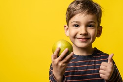 Small caucasian boy little child standing in front of yellow modern background holding green apple and thumbs up smiling - healthy eating concept with copy space