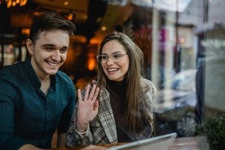 Two people young adult couple woman and man boyfriend and girlfriend sitting at cafe happy smile waving to say hello in day real people copy space relationship concept