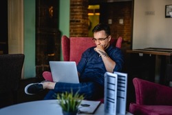 One man young caucasian male sitting in the office at home or at cafe working on laptop computer alone hold his chin thinking and contemplating while checking email or project real people copy space