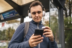 One man young adult male stand at public transport bus station waiting with headphones and mobile smart phone in winter or autumn day with backpack student or tourist city life copy space