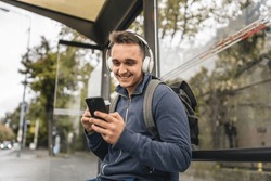 One man young adult male sit at public transport bus station waiting with headphones and mobile smart phone in winter or autumn day with backpack student or tourist city life copy space happy smile