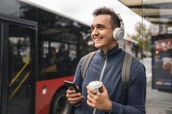 One man young adult male stand at public transport bus station taking bus with headphones and mobile smart phone in winter or autumn day with backpack student tourist city life copy space happy smile