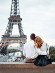 A young couple (a guy and a red-haired girl) in white shirts sit and hug against the backdrop of the Eiffel tower in Paris. Romantic trip to Europe.