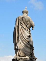 Statue of Saint Peter from the back on Vatican square with a bird sitting on its head. Blue sky with white clouds are in the background.