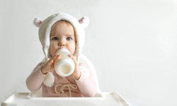 Little toddler girl in a warm fluffy hat drinks milk from a bottle while sitting. Half-length portrait. White gray background.