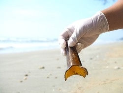 a rubber gloved hand picking up brown broken glass bottle background are sand beach sea and sky concept garbage on the beach
