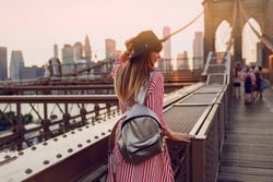 View from back of  traveling woman in red dress, stylish black  hat and silver bag pack enjoying amazing view from Brooklyn bridge in New York. Sunset colors.
