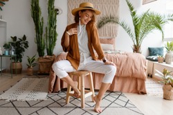 Cozy home atmosphere. Stylish woman in linen clothes sitting on chair over bed and home plants. Soft colors.
