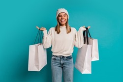 Studio winter portrait of  smiling blond woman in white hat and whool sweater  a holding  shopping bags ,posing in studio over turquose background. 