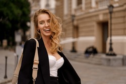 Beautiful smiling woman in elegant black suit  posing outdoor in old european city.  Blond wavy hairs, perfet skin.