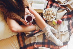 Soft cozy photo of slim tan woman in warm sweater on the bed with cup of tea in hands, top view point. Girl sitting on checkered plaid near old books and cookies.