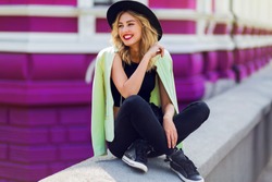 Outdoor lifestyle  close up portrait of happy blonde young woman in stylish  casual outfit sitting on bridge on the street. Pretty hipster girl having fun and enjoying  holidays. 