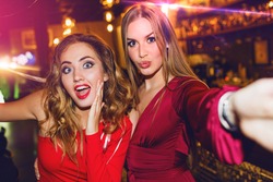 Two best friends blonde  women in evening casual red dress taking selfie at party, drinking cocktails, celebrating new year / birthday. Having fun, enjoying  holidays, funny grimaces ,flash portrait. 