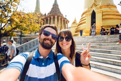 Successful traveling couple in love taking a selfie on phone  at Grand Palace temples in Bangkok  Pretty girl and her handsome boyfriend with beard having fun, crazy emotional faces , laughing.