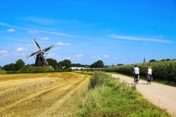 Cycling track with couple of two cyclists in rural flat dutch maas landscape with corn field, trees,   windmill (Molen de grauwe beer) against blue summer sky - Beesel, Netherlands