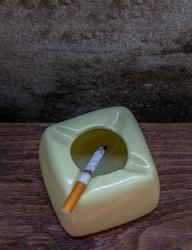 Cigarette that were burning placed on greyish green ashtray on old wooden rustic table. Copy space, Selective focus.