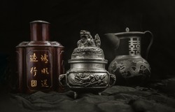 Silver antique incense burner, Chinese antique tea leaf iron storage jar (Characters chinese is Name of the tea) and Chinese antique teapot (Characters chinese is Double Happiness) on dark background.