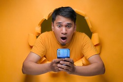 Portrait Young Asian man through torn yellow paper hole, stonished looking at smartphone screen in shock with mouth round because of deep astonishment and shock of great news