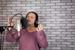 Portrait of male musician who sings song into microphone and plays guitar while sitting on gray background. Emotional middle-aged man dressed in rock style sings author's song. Studio shot.