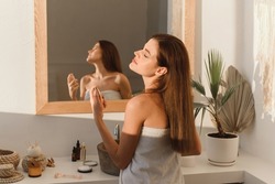 A beautiful young woman sprays perfume on her neck while standing in the bathroom.