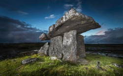 Ancient portal thomb Poulnabrone Dolmen standing on green grass in a rocky field in Glenslane Ireland lighted by flashlight before sunset with a background of blue and orange sky
